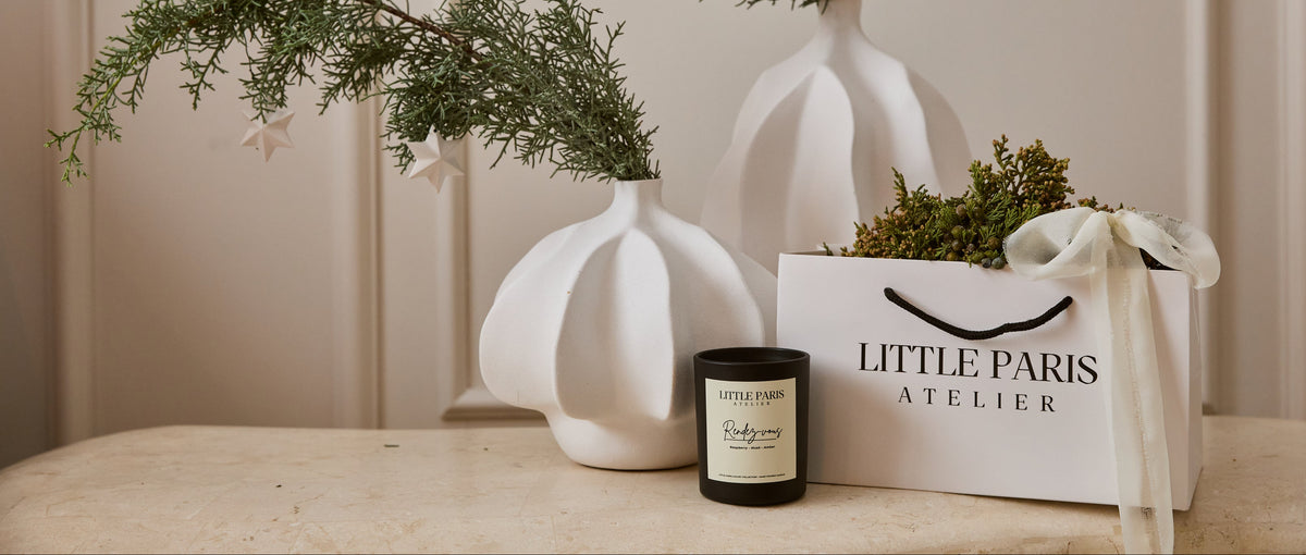 Lumanare Little Paris langa vase albe din ceramica si o punga de cadou, in lumina calda de dupa-amiaza